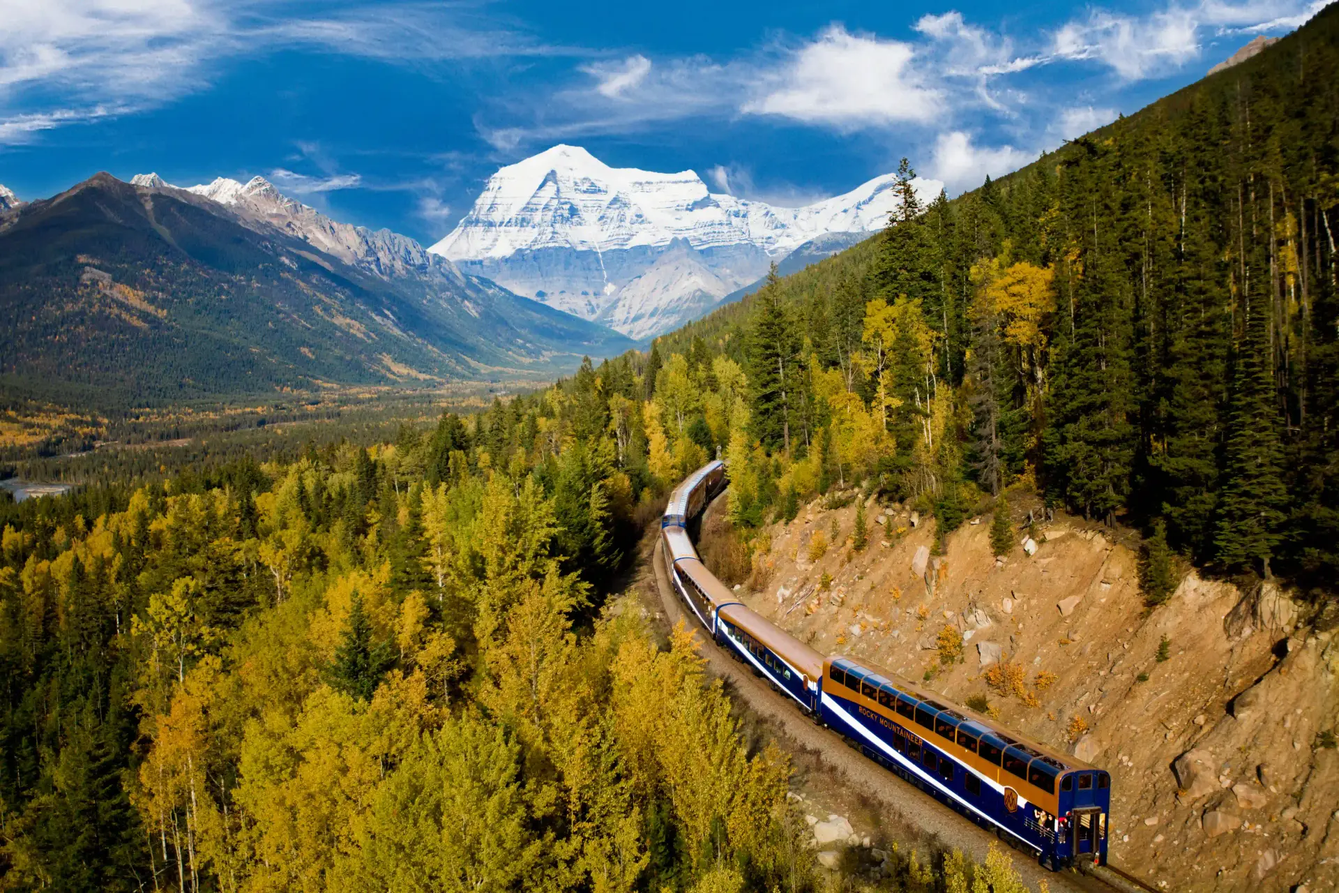Scenic view of a train traveling through vibrant autumn forests with majestic mountains in the background, showcasing the beauty of nature in a mountainous region.