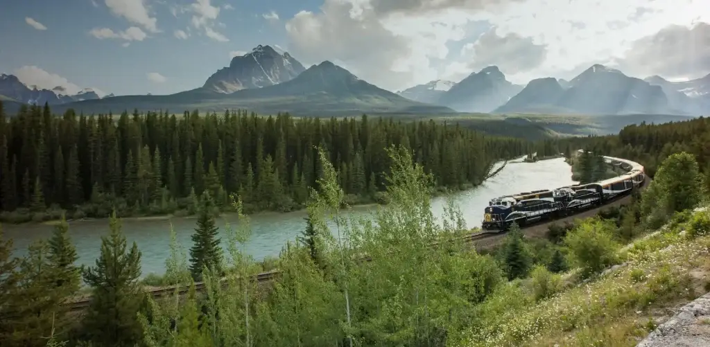 Scenic view of a train traveling along a river surrounded by lush forests and majestic mountains under a partly cloudy sky.