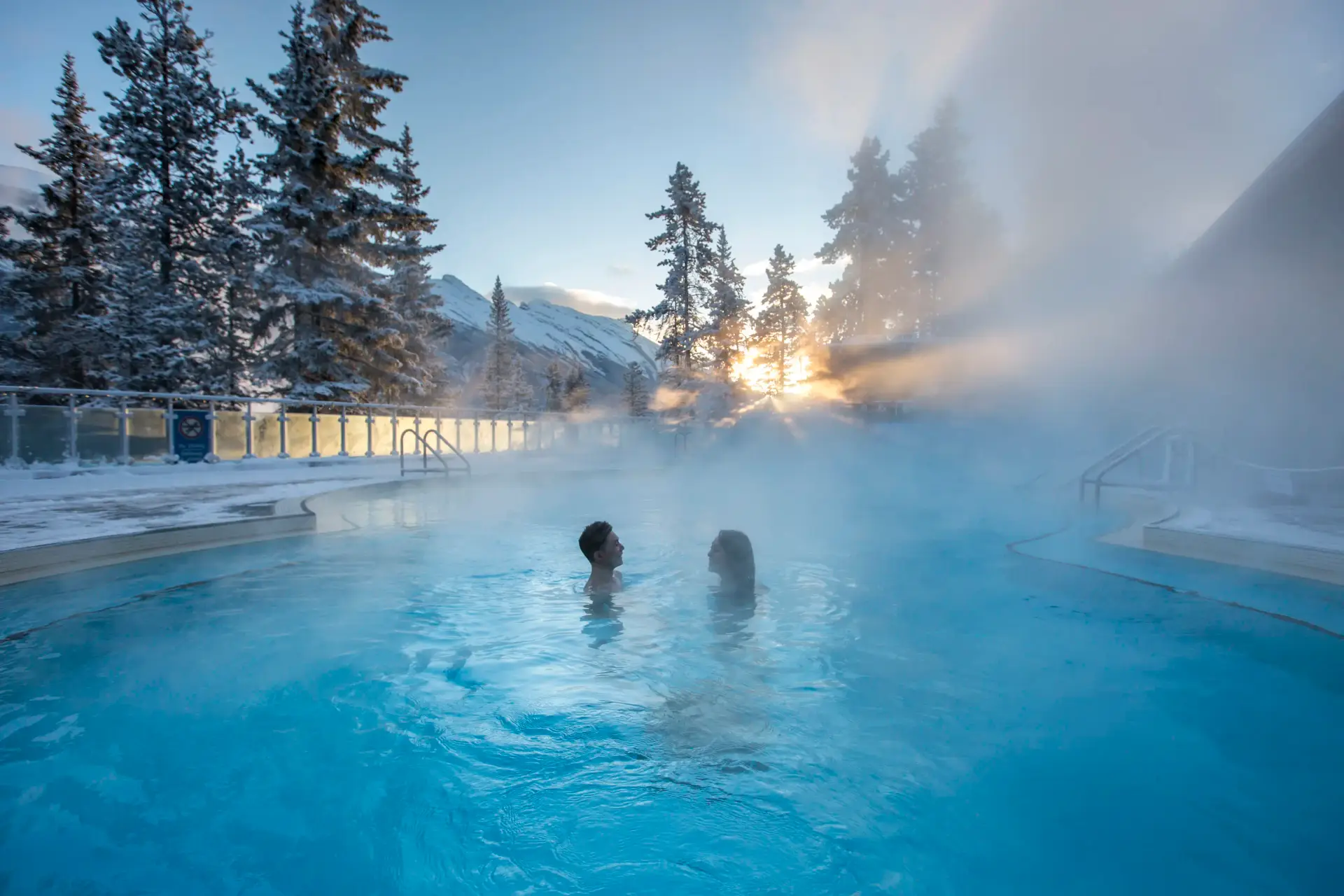 Couple enjoying a warm soak in a steaming outdoor hot spring pool surrounded by snowy trees and mountains during sunrise.
