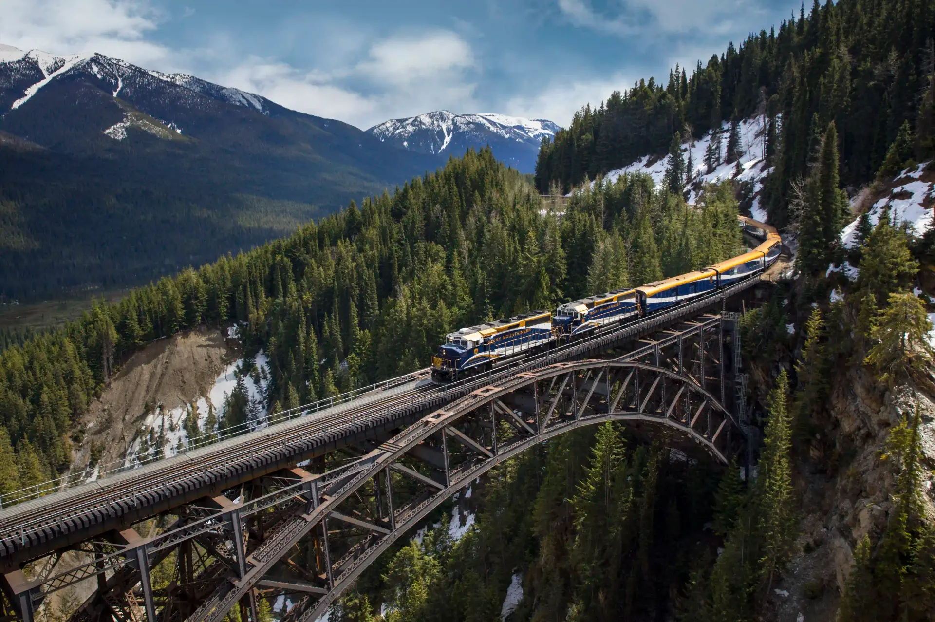 A scenic view of a train crossing a wooden trestle bridge surrounded by lush green forests and snow-capped mountains, showcasing the beauty of nature and railway travel.