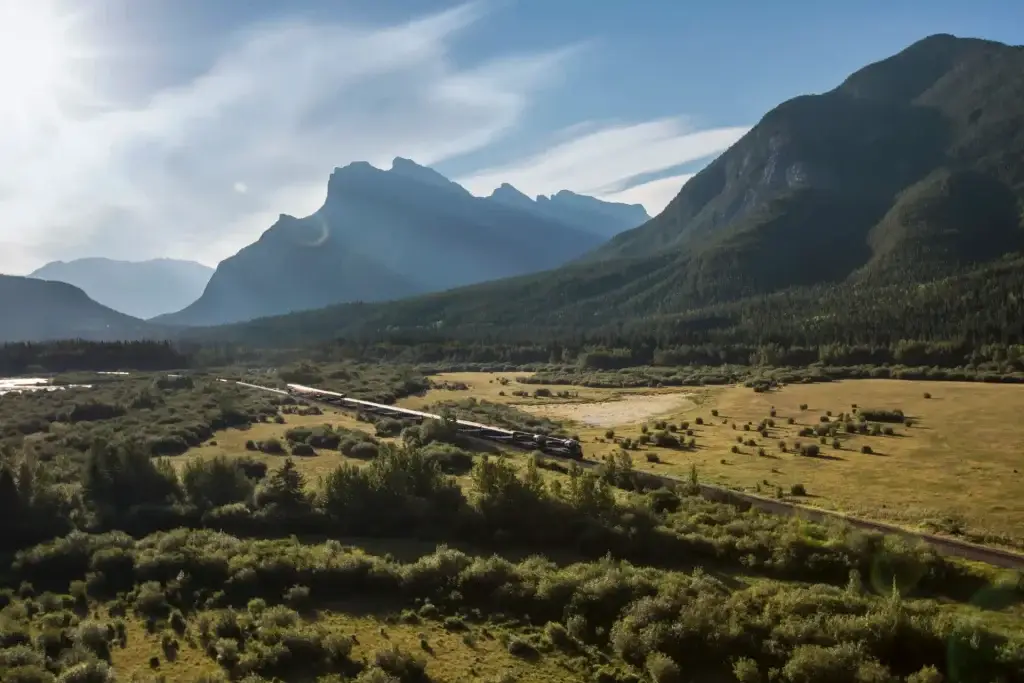 Scenic view of a train traveling through a lush valley surrounded by majestic mountains and blue skies, showcasing the natural beauty of the landscape.