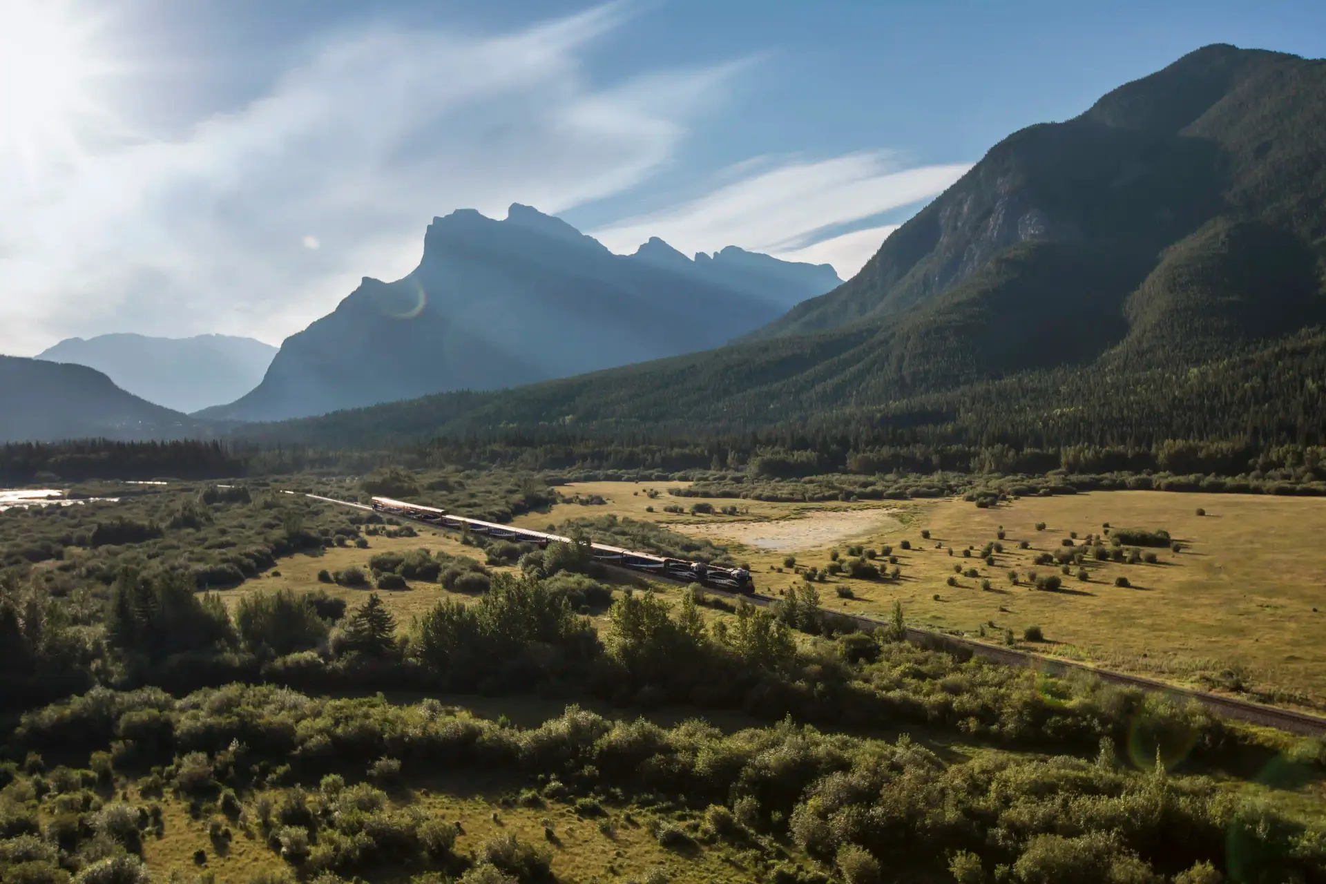 Scenic view of a train traveling through a lush valley surrounded by majestic mountains and blue skies, showcasing the natural beauty of the landscape.