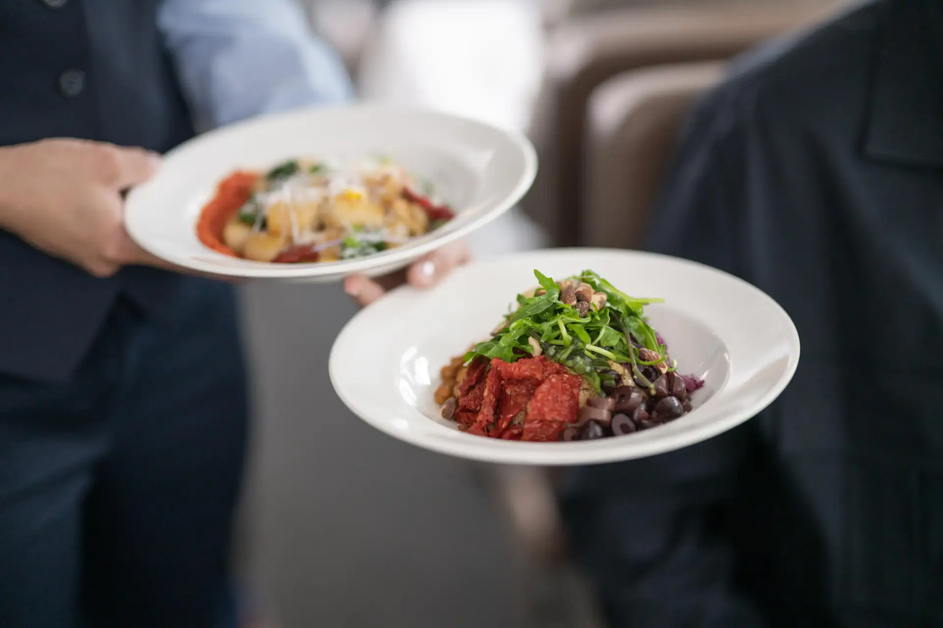 A waiter serving two plates of colorful gourmet dishes, one featuring pasta with vegetables and cheese, and the other a vibrant salad with greens, beans, and sun-dried tomatoes, in a modern dining setting.