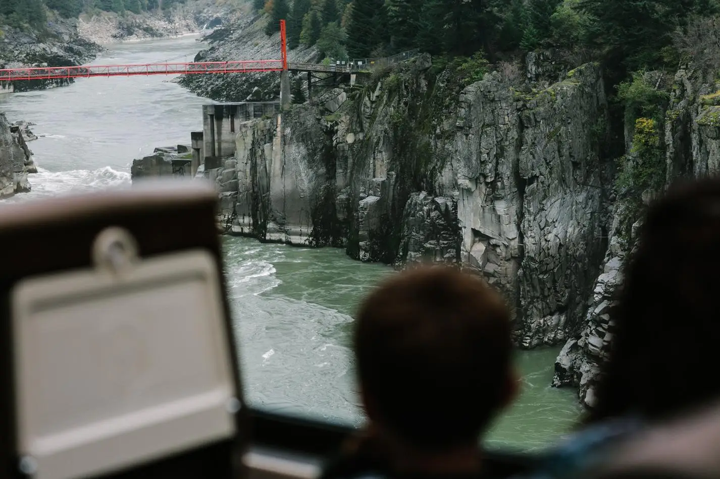 A scenic view of a river surrounded by rocky cliffs, with a red bridge in the background, as seen from the perspective of two individuals. The image captures the natural beauty and rugged landscape, highlighting the contrast between the flowing water and the rocky terrain.