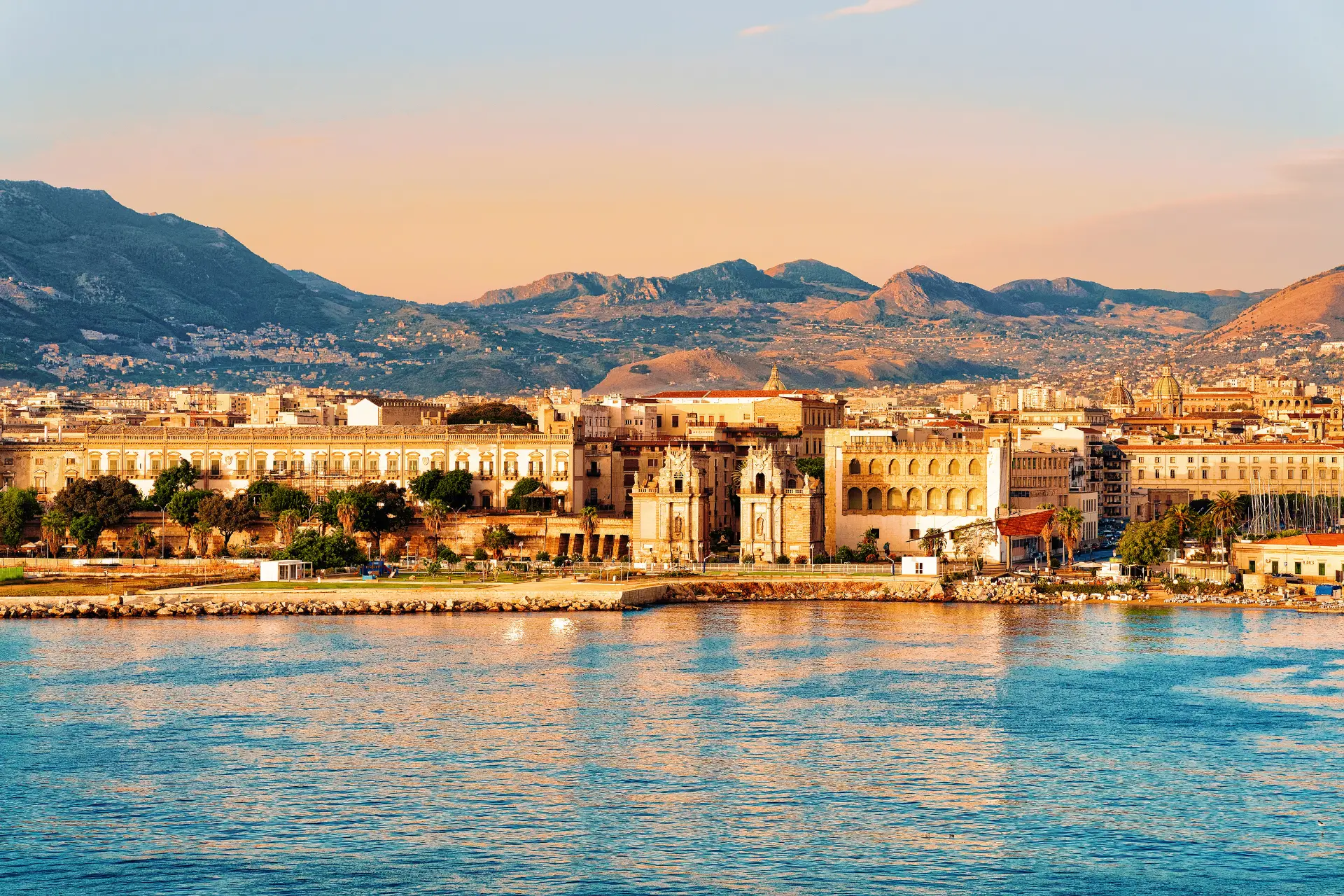 Scenic view of Palermo, Italy, showcasing historical architecture along the coastline with mountains in the background during sunset. The image captures the vibrant colors of the sky reflected in the water, highlighting Palermo's blend of natural beauty and urban landscape.