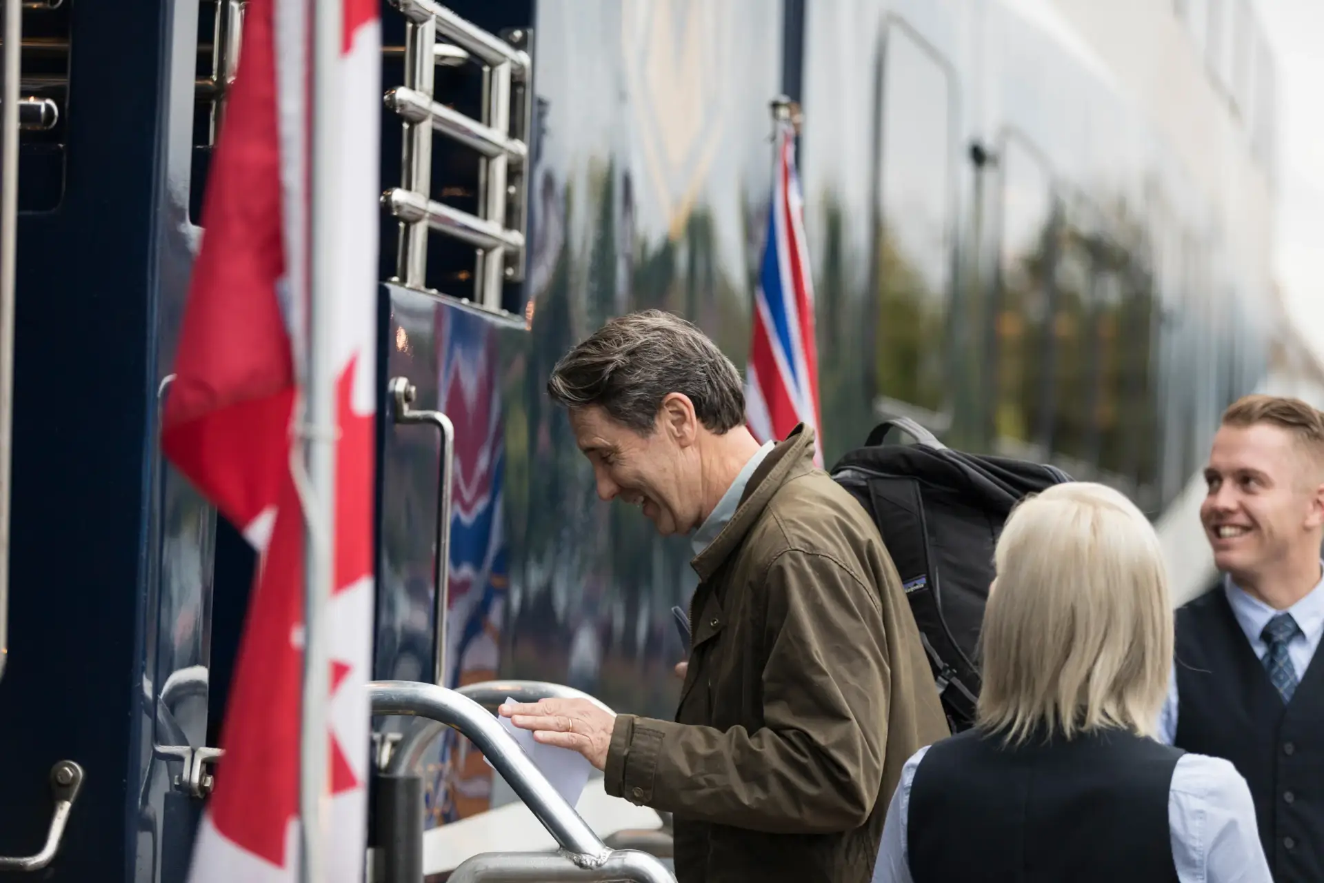 Passengers boarding a luxury train, smiling and engaging with staff, with Canadian and British flags in the background, showcasing a travel experience.