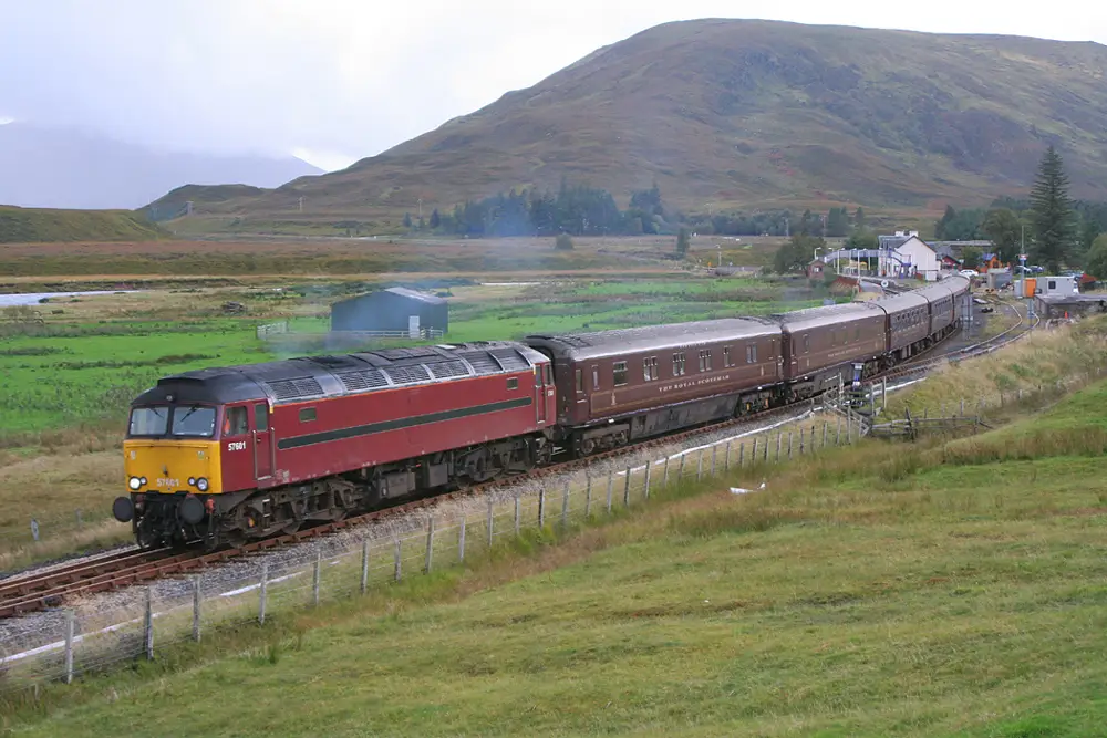 A diesel locomotive traveling along a scenic railway in a rural landscape, surrounded by green fields and distant hills, with a vintage passenger train in tow.