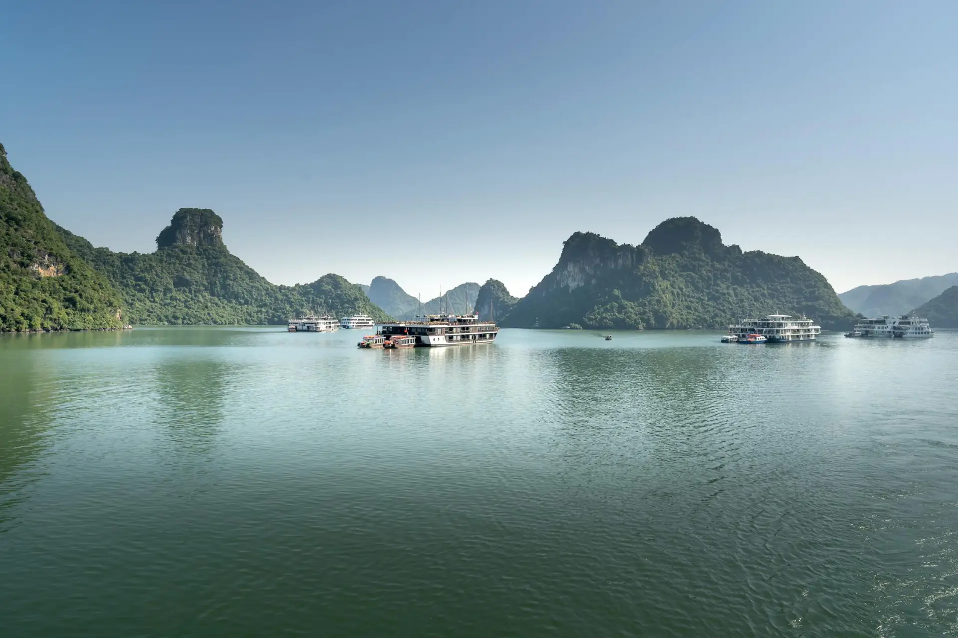 A serene view of Ha Long Bay featuring multiple boats anchored amidst lush green mountains under a clear blue sky, showcasing the natural beauty and tranquility of this UNESCO World Heritage site in Vietnam.