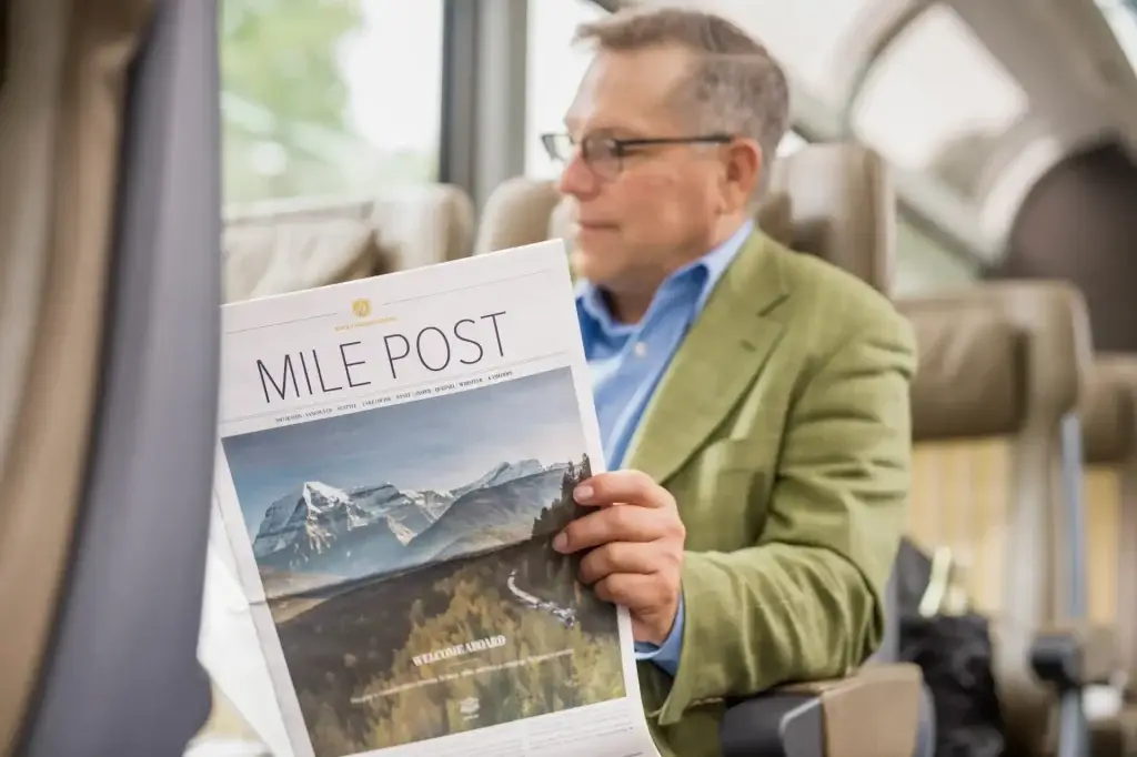 A man in a green blazer reading the "Mile Post" newspaper while seated on a train, with scenic mountain views visible through the window. The image captures the relaxed atmosphere of train travel and exploration.