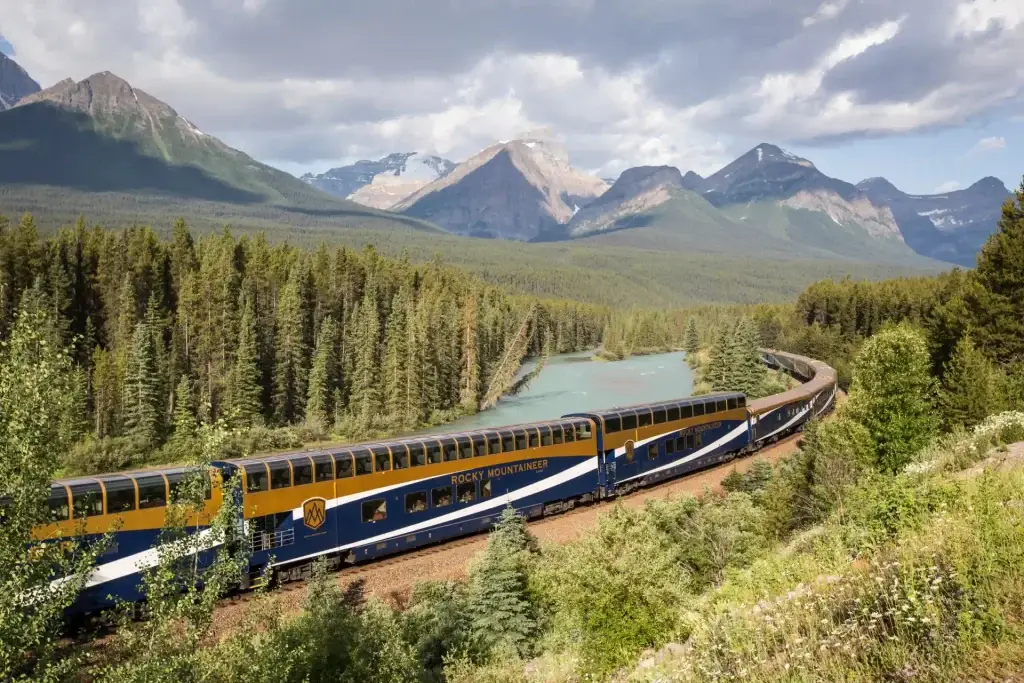 Scenic view of the Rocky Mountaineer train traveling through lush forests and mountains, with a river winding alongside the tracks under a partly cloudy sky.