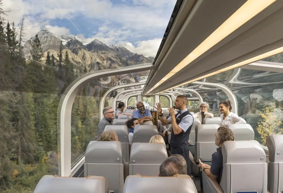 Interior view of a scenic train journey featuring passengers enjoying the panoramic views through large glass windows, with a conductor providing a guided experience amidst a backdrop of majestic mountains and lush forests.