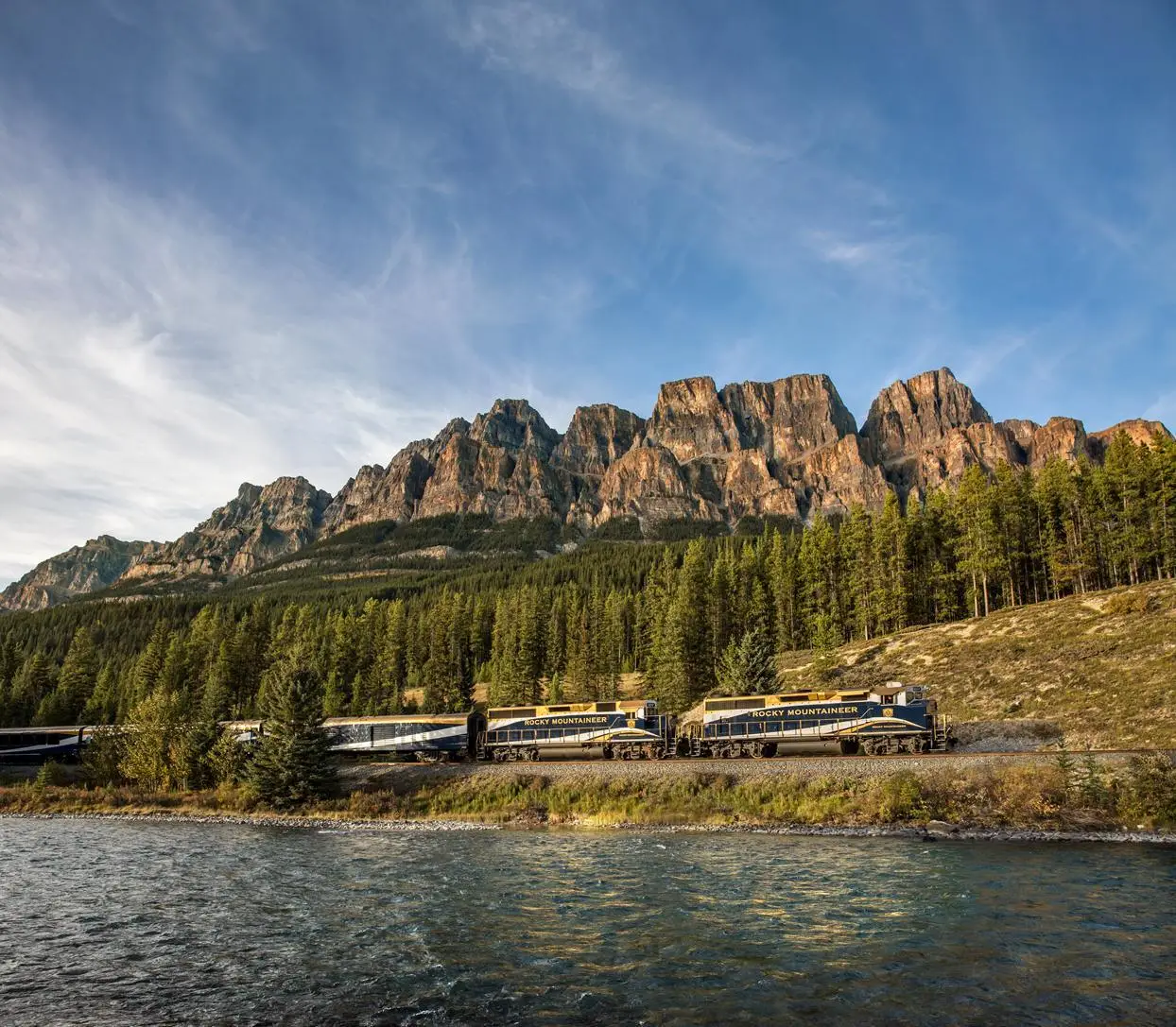 Scenic view of the Rocky Mountaineer train traveling along a river, with majestic mountain peaks and lush forests in the background, showcasing the breathtaking landscape of the Canadian Rockies.