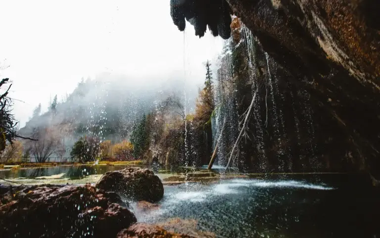 Waterfall cascading over rocky terrain into a serene pool, surrounded by misty mountains and autumn foliage, creating a tranquil natural landscape.