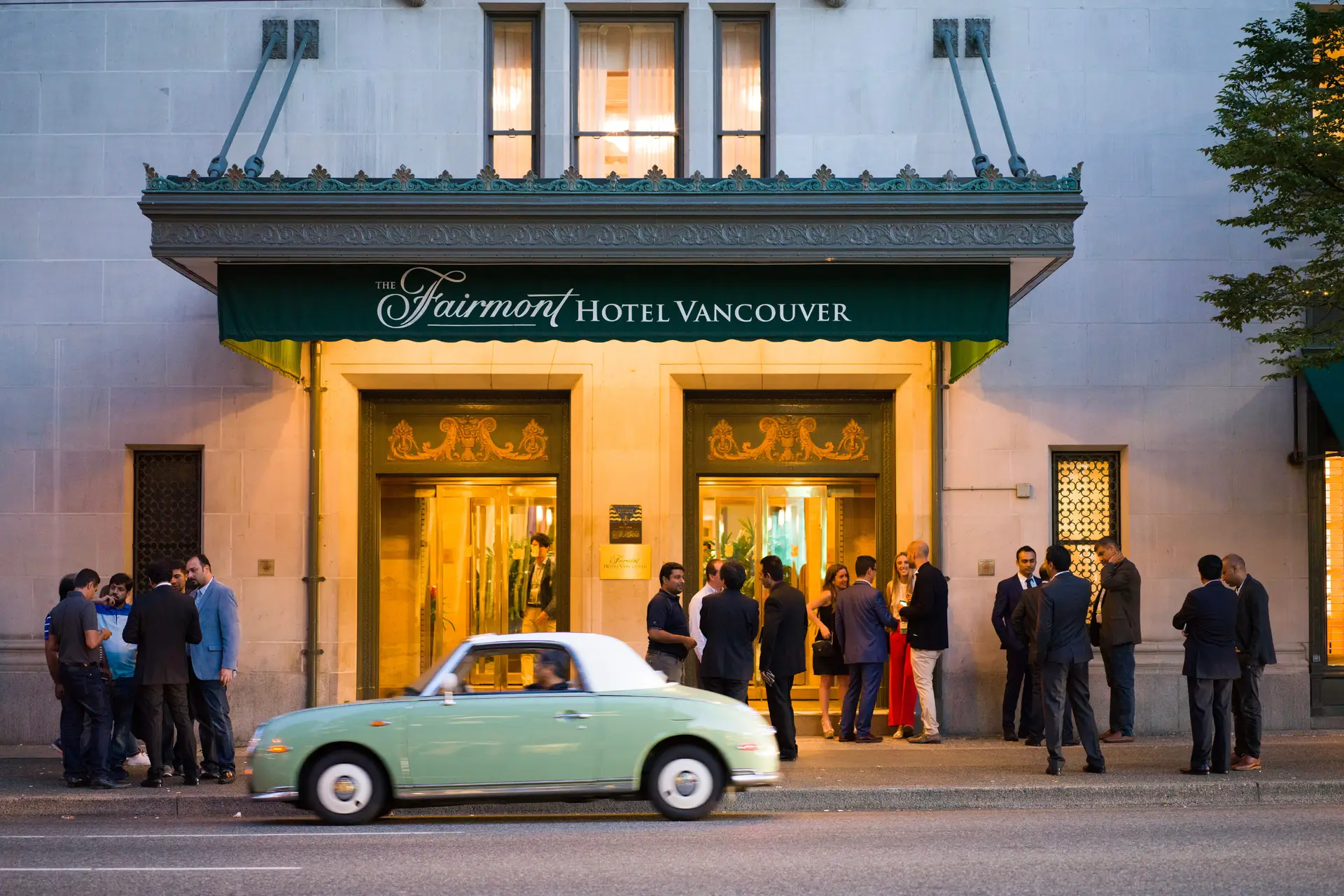 Exterior view of the Fairmont Hotel Vancouver with a group of people socializing at the entrance, showcasing the hotel's elegant architecture and a vintage car driving by.