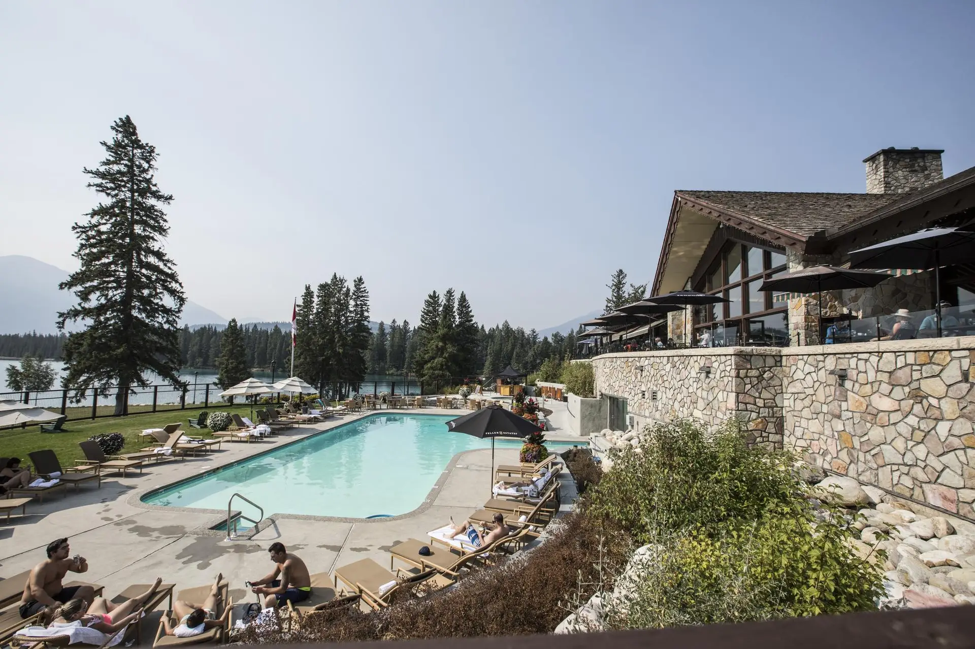 Scenic outdoor swimming pool at a lakeside resort, surrounded by lounge chairs and umbrellas, with a modern stone building and lush trees in the background, ideal for relaxation and leisure activities.