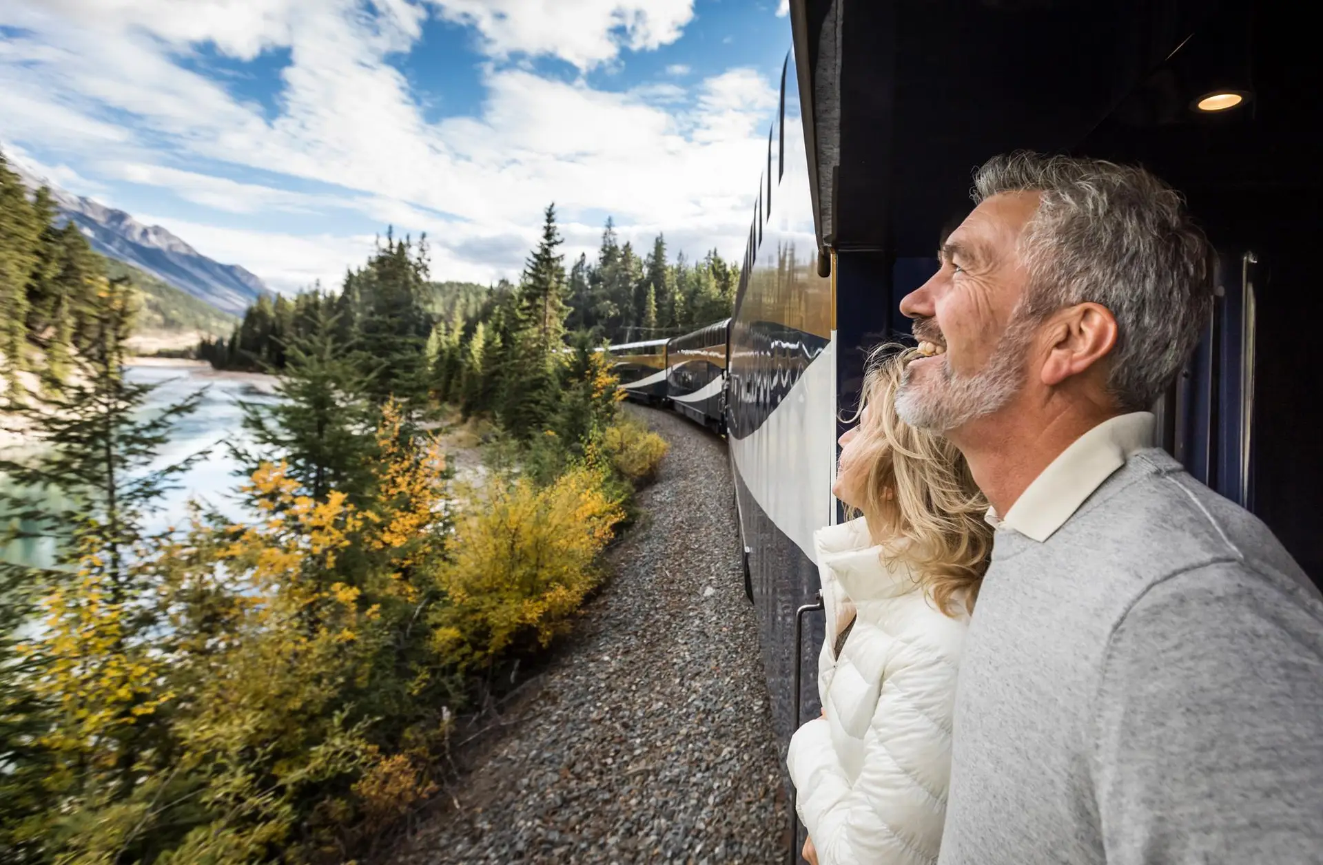 Couple enjoying scenic views from a train window, surrounded by lush forests and a river, with mountains in the background and a bright blue sky. Perfect for travel and adventure enthusiasts.