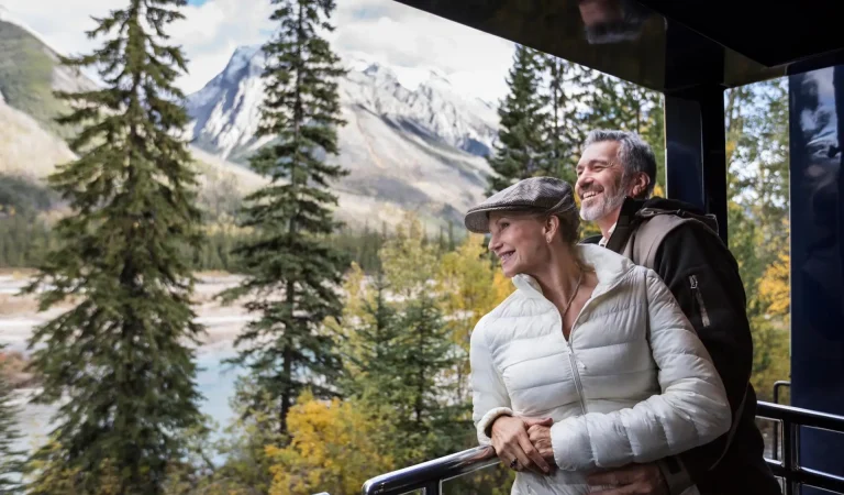 Couple enjoying a scenic view of mountains and trees from a balcony, dressed in warm clothing, surrounded by autumn foliage and a river.