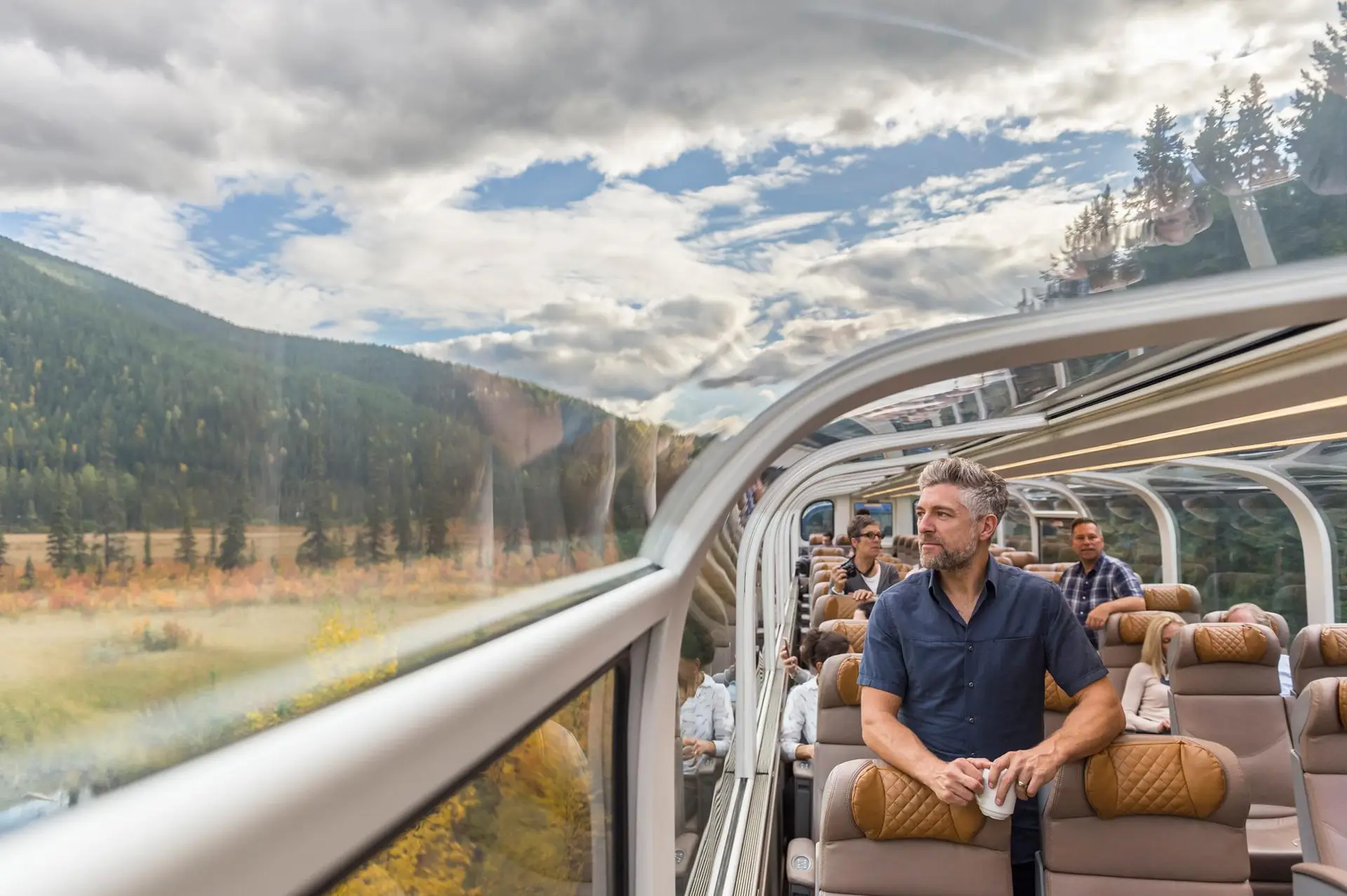 Interior view of a scenic train journey, featuring passengers enjoying panoramic mountain views through large glass windows while seated in comfortable, modern train seats.