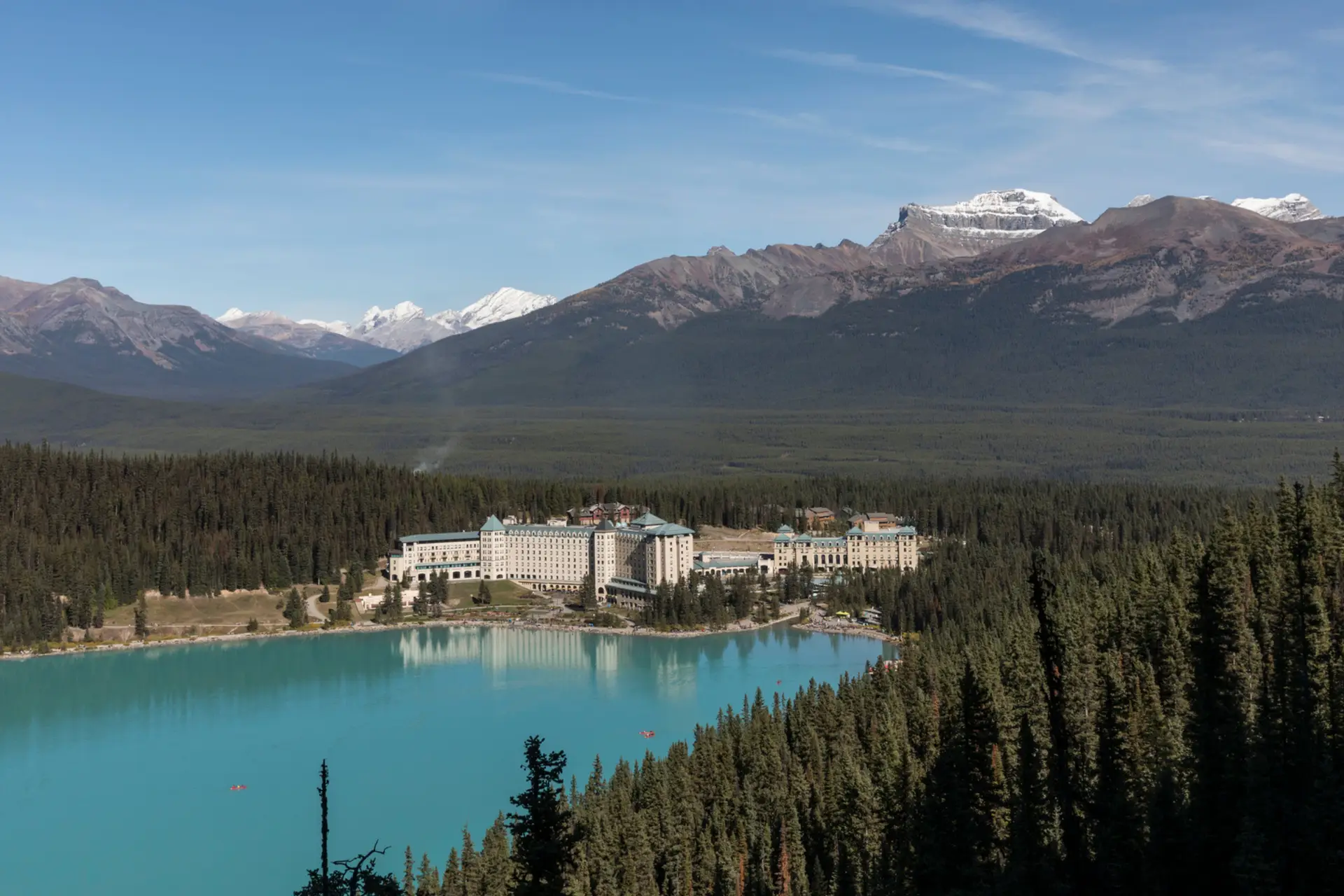 Scenic view of a luxury hotel nestled by a turquoise lake, surrounded by dense forests and majestic mountains in the background, showcasing the natural beauty of the landscape.