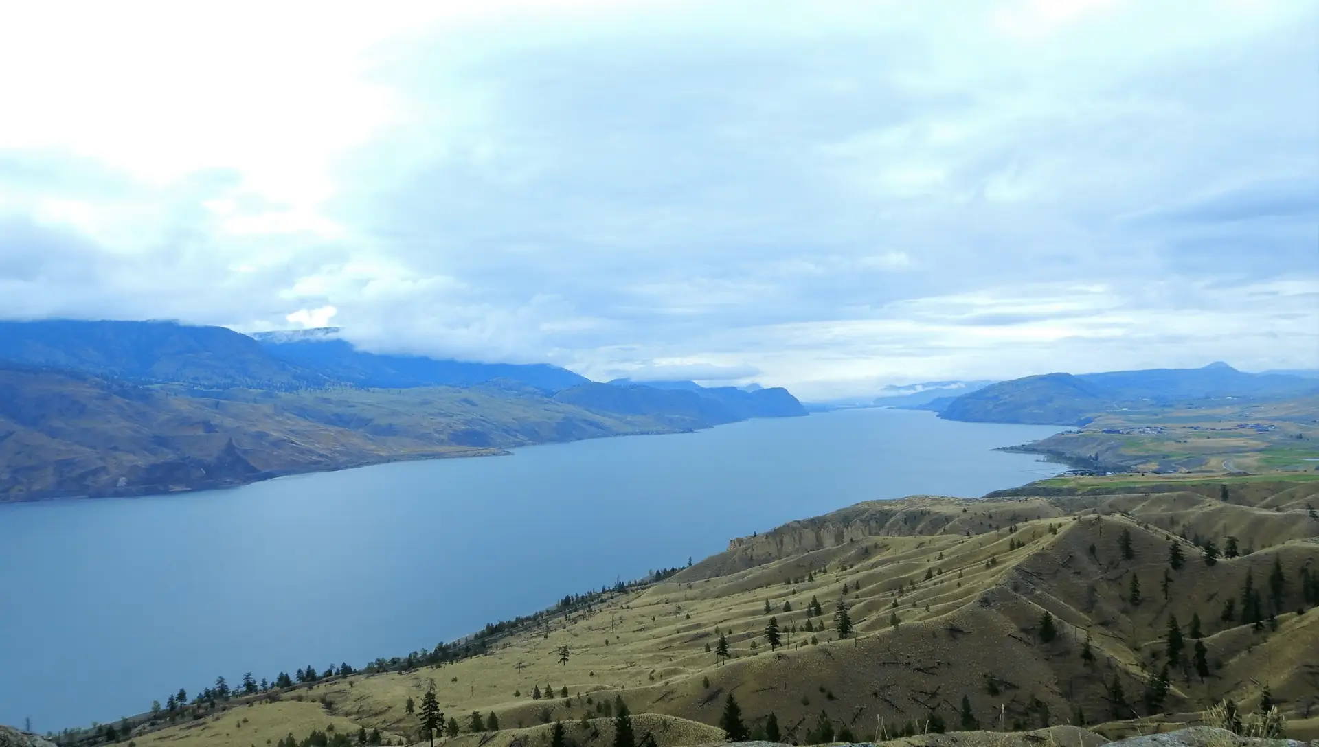 Panoramic view of a serene lake surrounded by rolling hills and mountains under a cloudy sky, showcasing the natural beauty of the landscape. Ideal for nature enthusiasts and outdoor exploration.