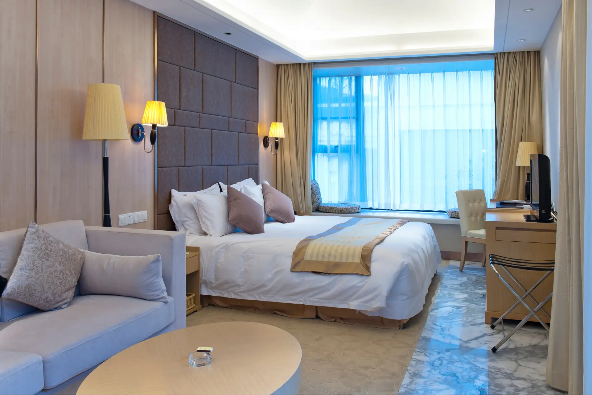 Modern hotel room featuring a king-size bed with decorative pillows, a cozy seating area, and large windows with sheer curtains. The room is elegantly designed with wooden paneling, stylish lamps, and a marble floor, providing a comfortable and inviting atmosphere for guests.