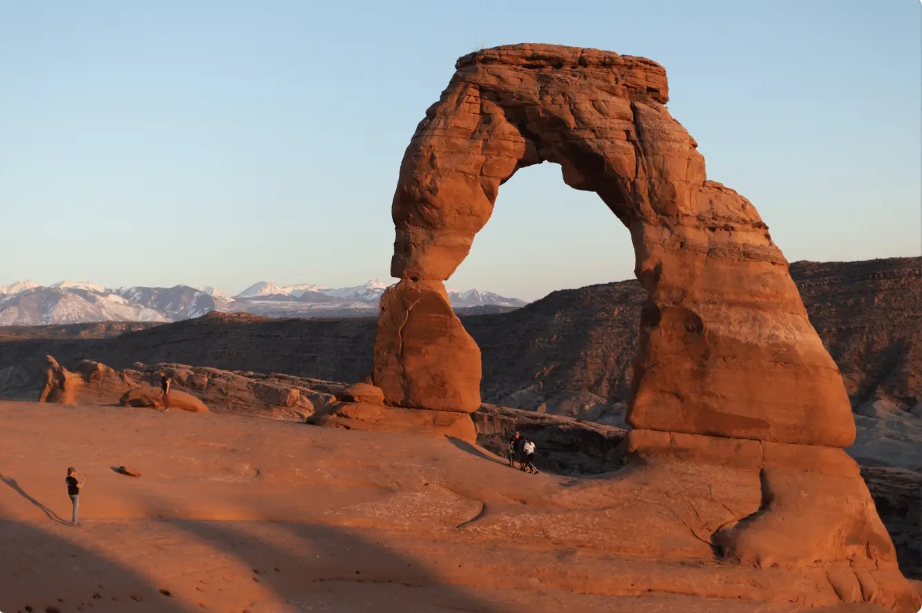 Delicate Arch at Arches National Park, Utah, showcasing its iconic natural sandstone arch against a backdrop of snow-capped mountains during sunset. Two visitors are seen exploring the area, emphasizing the scale of the arch.