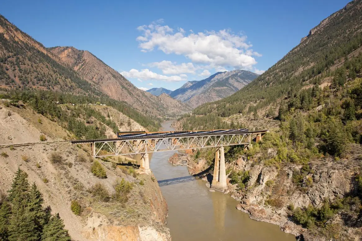 A scenic view of a train crossing a steel bridge over a river, surrounded by mountains and lush greenery, under a clear blue sky.