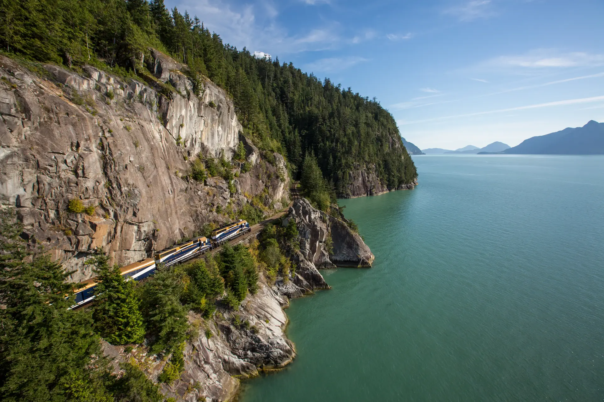 Scenic view of a train traveling along a rocky coastline surrounded by lush green forests and calm turquoise waters, under a clear blue sky. Ideal for showcasing natural beauty and rail travel in picturesque landscapes.