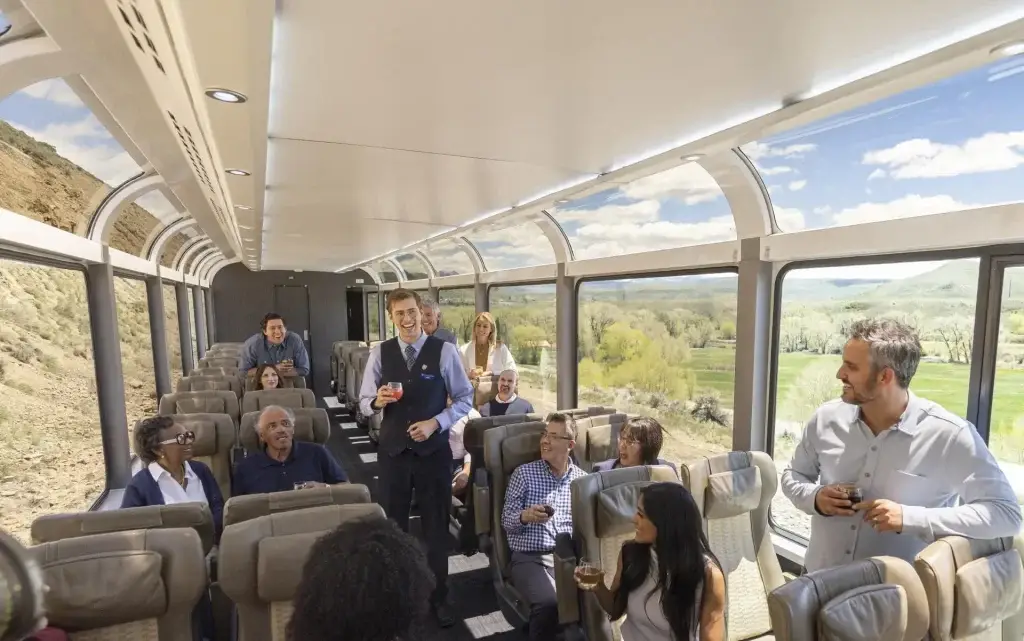 Interior view of a luxurious train carriage with passengers enjoying drinks and socializing, featuring large windows that showcase scenic landscapes. A train attendant engages with guests, enhancing the travel experience.