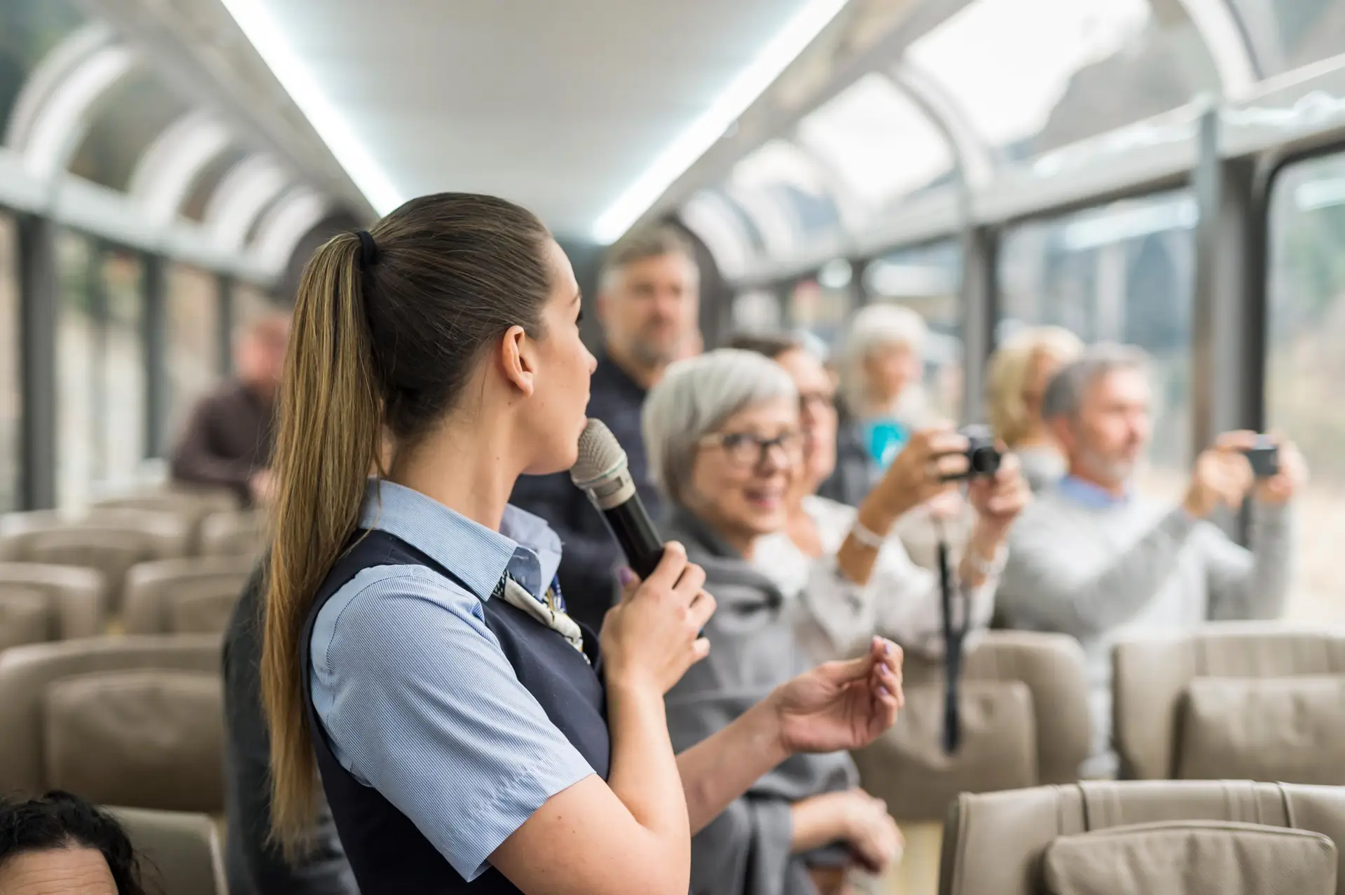 Tour guide speaking to passengers in a scenic train car, with travelers capturing moments on cameras; a lively atmosphere showcasing tourism and guided experiences.