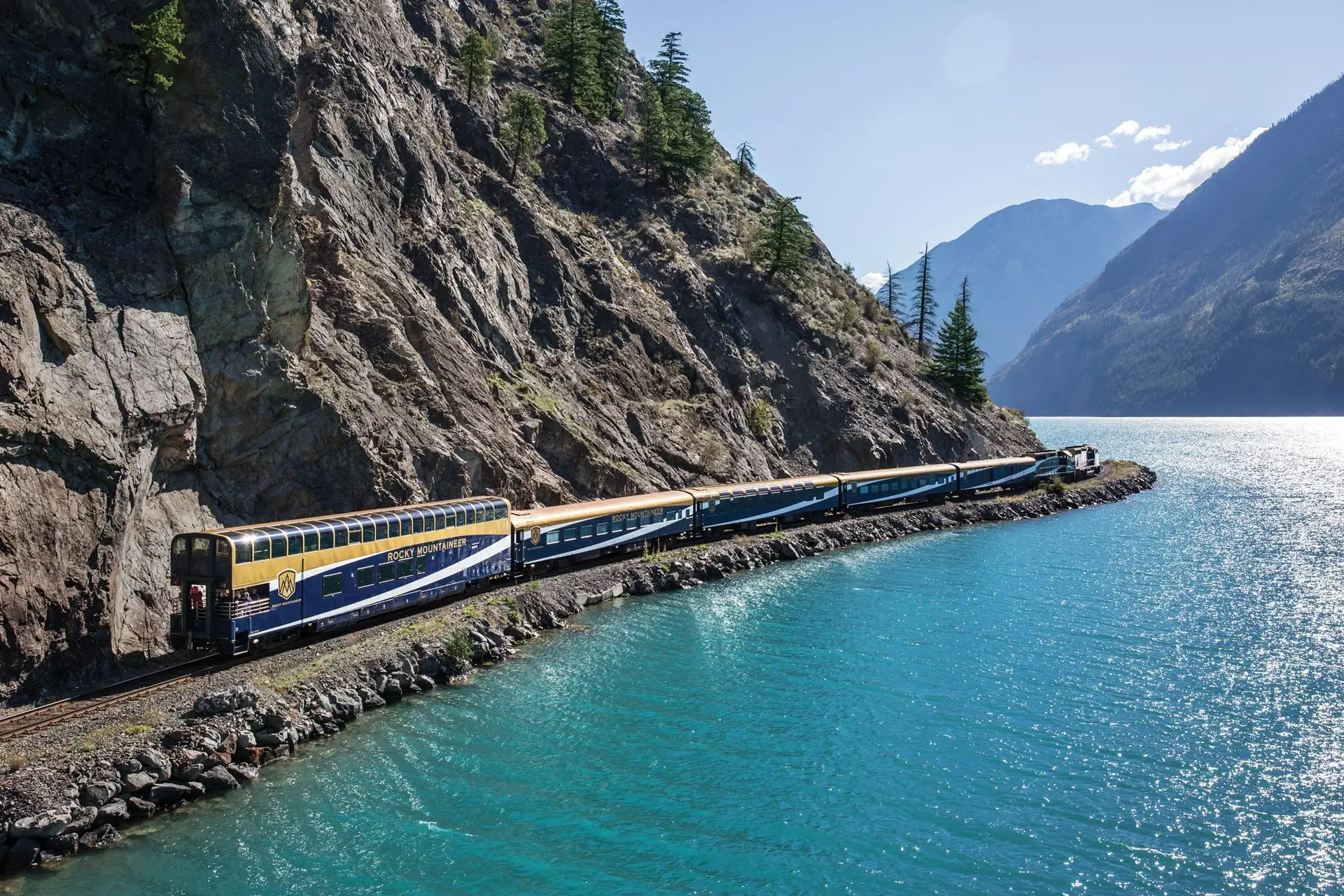 Scenic view of a Rocky Mountaineer train traveling along a rocky shoreline beside turquoise waters, surrounded by mountains and evergreen trees in a bright, sunny landscape.