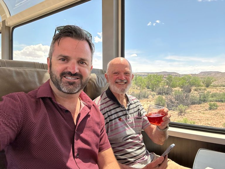 Two men enjoying a scenic train ride, smiling as they relax in comfortable seating next to a large window with a view of the natural landscape. One man is holding a glass of red drink while the other is taking a selfie.