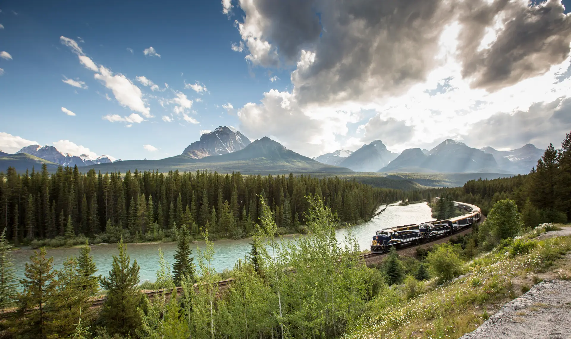 Scenic view of a train traveling along a river surrounded by lush forests and majestic mountains under a partly cloudy sky in a picturesque landscape.