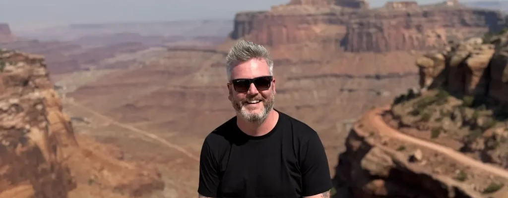 Man with gray hair and sunglasses smiles while sitting on a rock ledge, with the breathtaking landscape of canyons and mountains in the background under a clear blue sky.
