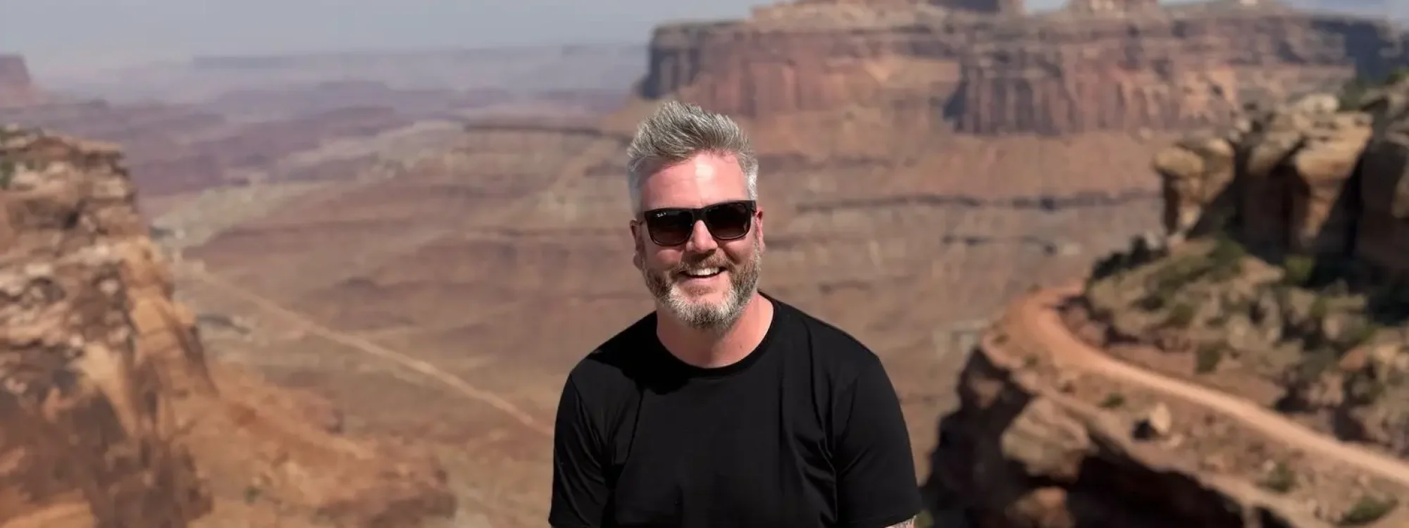 Man with gray hair and sunglasses smiles while sitting on a rock ledge, with the breathtaking landscape of canyons and mountains in the background under a clear blue sky.