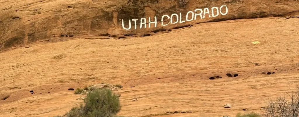 Rocky landscape featuring the inscription "UTAH COLORADO" on a sandstone cliff, surrounded by sparse vegetation and rocky terrain typical of the region.