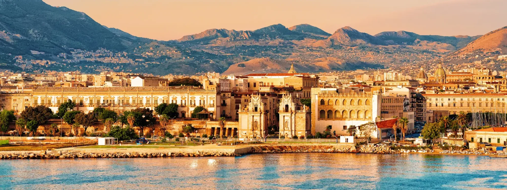 Scenic view of Palermo, Italy, showcasing historical architecture along the coastline with mountains in the background during sunset. The image captures the vibrant colors of the sky reflected in the water, highlighting Palermo's blend of natural beauty and urban landscape.