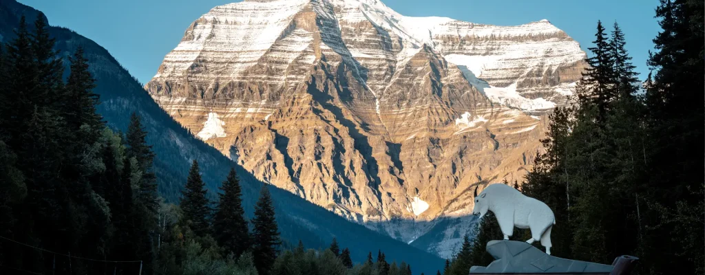 Mount Robson, the highest peak in the Canadian Rockies, towers majestically against a clear blue sky, showcasing snow-capped summits and rugged cliffs. In the foreground, a sign with a white mountain goat sculpture marks the entrance to Mount Robson Provincial Park, surrounded by dense evergreen forests and a winding road.