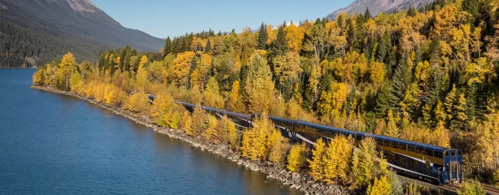 Scenic view of a train traveling along a lake, bordered by vibrant autumn foliage and mountains in the background, showcasing the beauty of fall colors and nature.