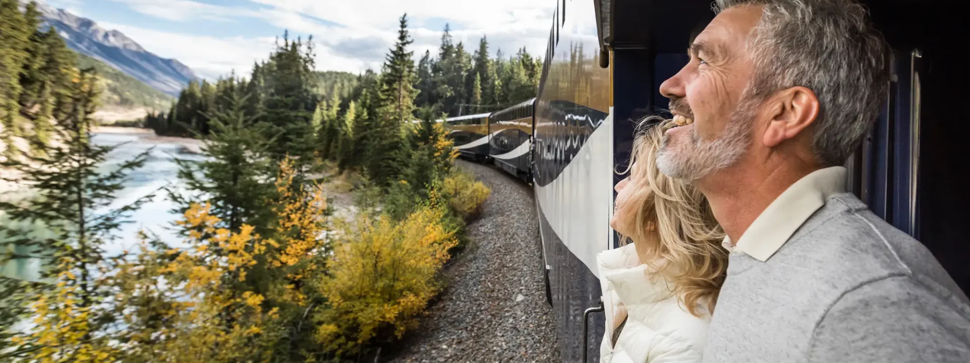 Couple enjoying scenic views from a train, surrounded by lush green forests and mountains under a bright blue sky, highlighting the beauty of nature during travel.