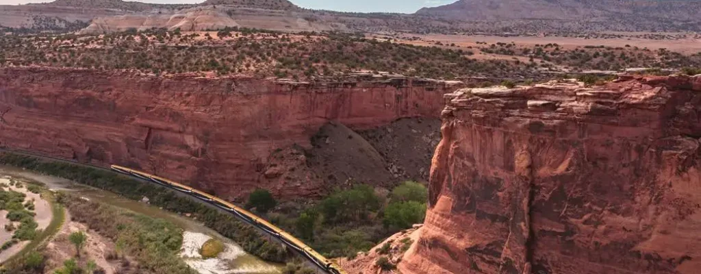 A scenic view of a winding train traveling along a riverbank, surrounded by stunning red rock cliffs and desert landscape under a blue sky with clouds. The image captures the natural beauty and geological features of the area, highlighting the harmony between the train and its picturesque environment.