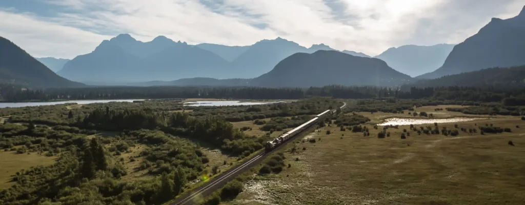 A scenic view of a train traveling along a railway through a lush green landscape, surrounded by mountains and a tranquil lake under a partly cloudy sky.