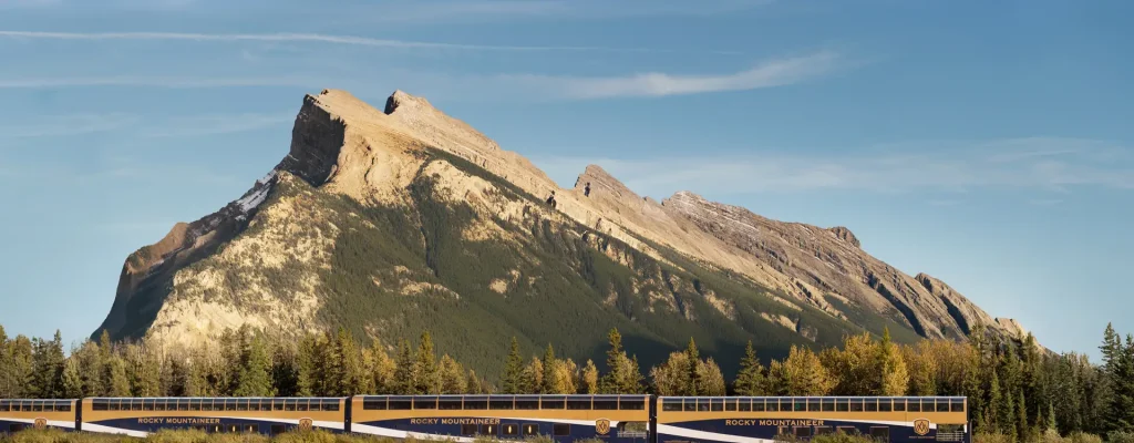 Scenic view of the Rocky Mountaineer train traveling through the picturesque landscape of the Canadian Rockies, featuring a prominent mountain peak against a clear blue sky and a reflective lake surrounded by lush greenery.
