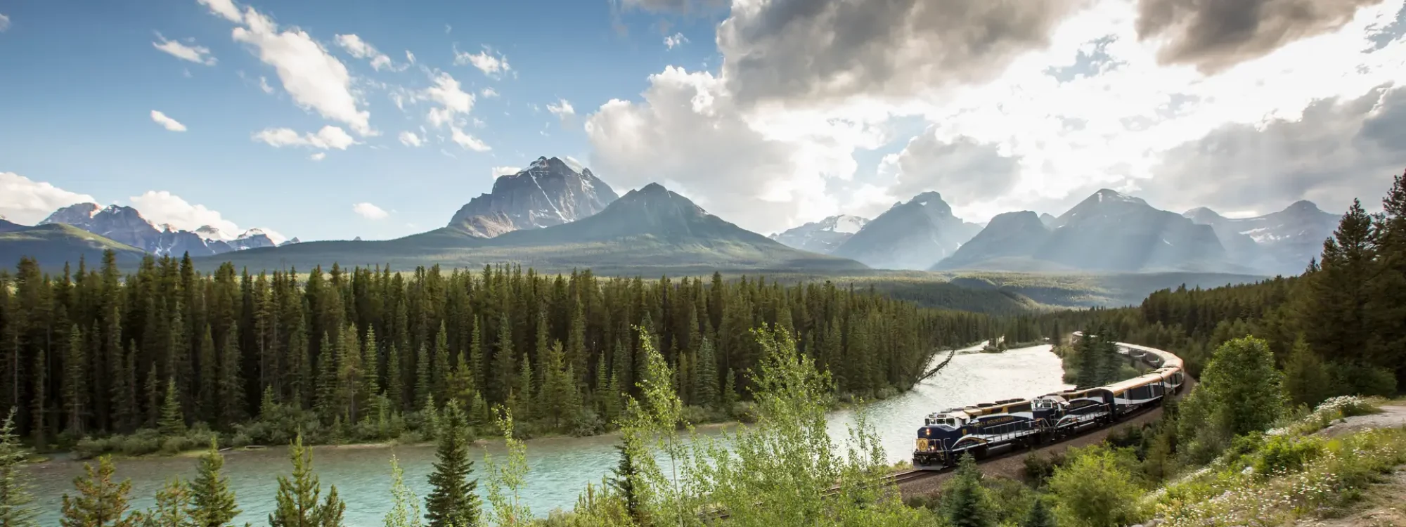 Scenic view of a train traveling along a river surrounded by lush forests and majestic mountains under a partly cloudy sky in a picturesque landscape.