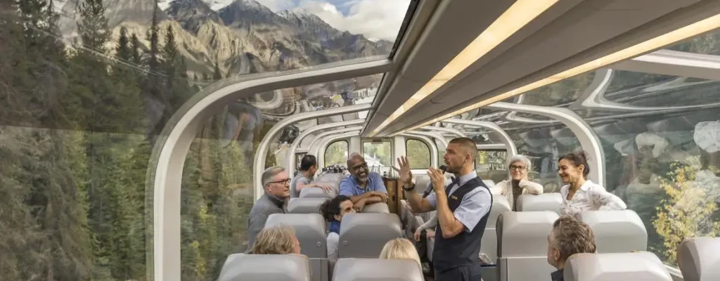 Interior view of a scenic train journey featuring passengers enjoying the panoramic views through large glass windows, with a conductor providing a guided experience amidst a backdrop of majestic mountains and lush forests.