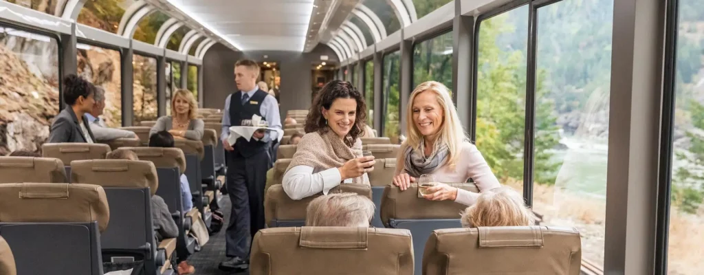 Interior view of a scenic train journey, featuring passengers enjoying drinks and engaging in conversation while a staff member serves refreshments. The large windows offer a picturesque view of nature outside, enhancing the travel experience.