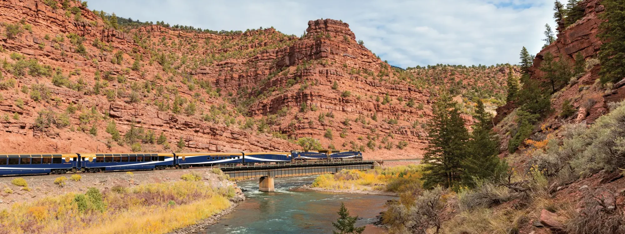 Scenic view of a train traveling along a riverbank, surrounded by red rock formations and lush greenery in a mountainous landscape. The image captures the natural beauty of the area, highlighting the contrast between the vibrant blue train and the earthy tones of the terrain.