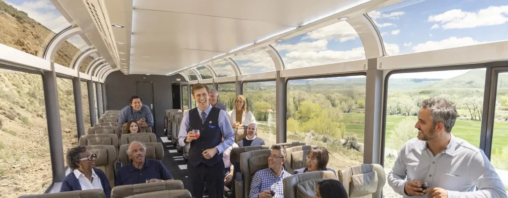 Interior view of a luxurious train carriage with passengers enjoying drinks and socializing, featuring large windows that showcase scenic landscapes. A train attendant engages with guests, enhancing the travel experience.